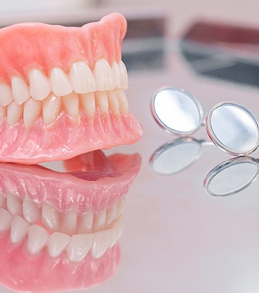Complete set of dentures next to dental mirror on reflective surface.