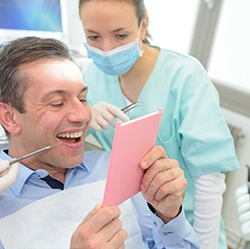 Man in blue shirt in dental chair smiling at reflection in hand mirror